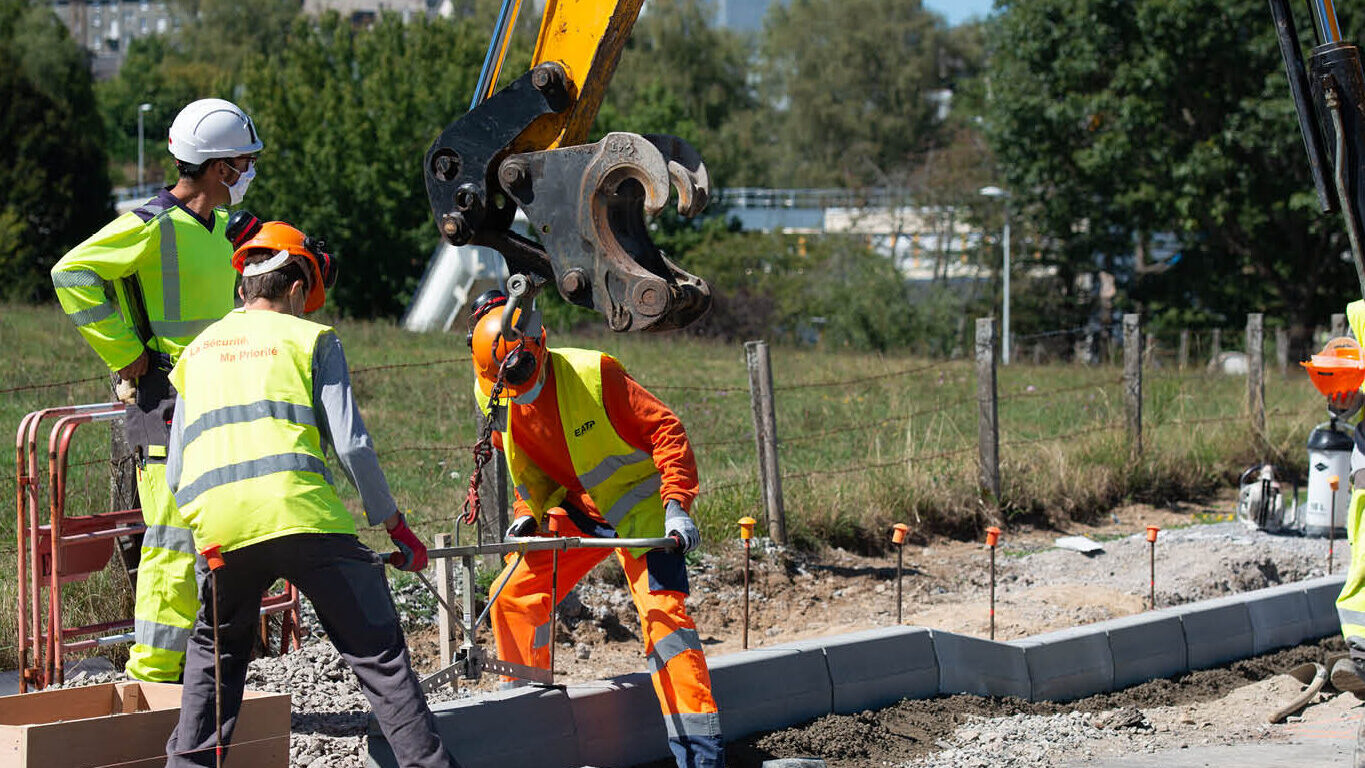 Une photo de chantier de pose de bordures. Avec 3 ouvriers qui pose la bordure et une pelle hydraulique qui manutentionne la bordure.